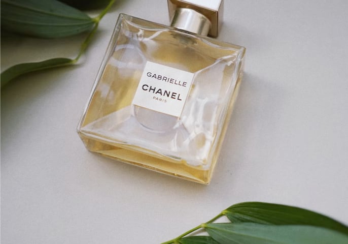 A bottle of Gabrielle Essence Eau De Parfum lying on a white table next to two branches with dark green leaves